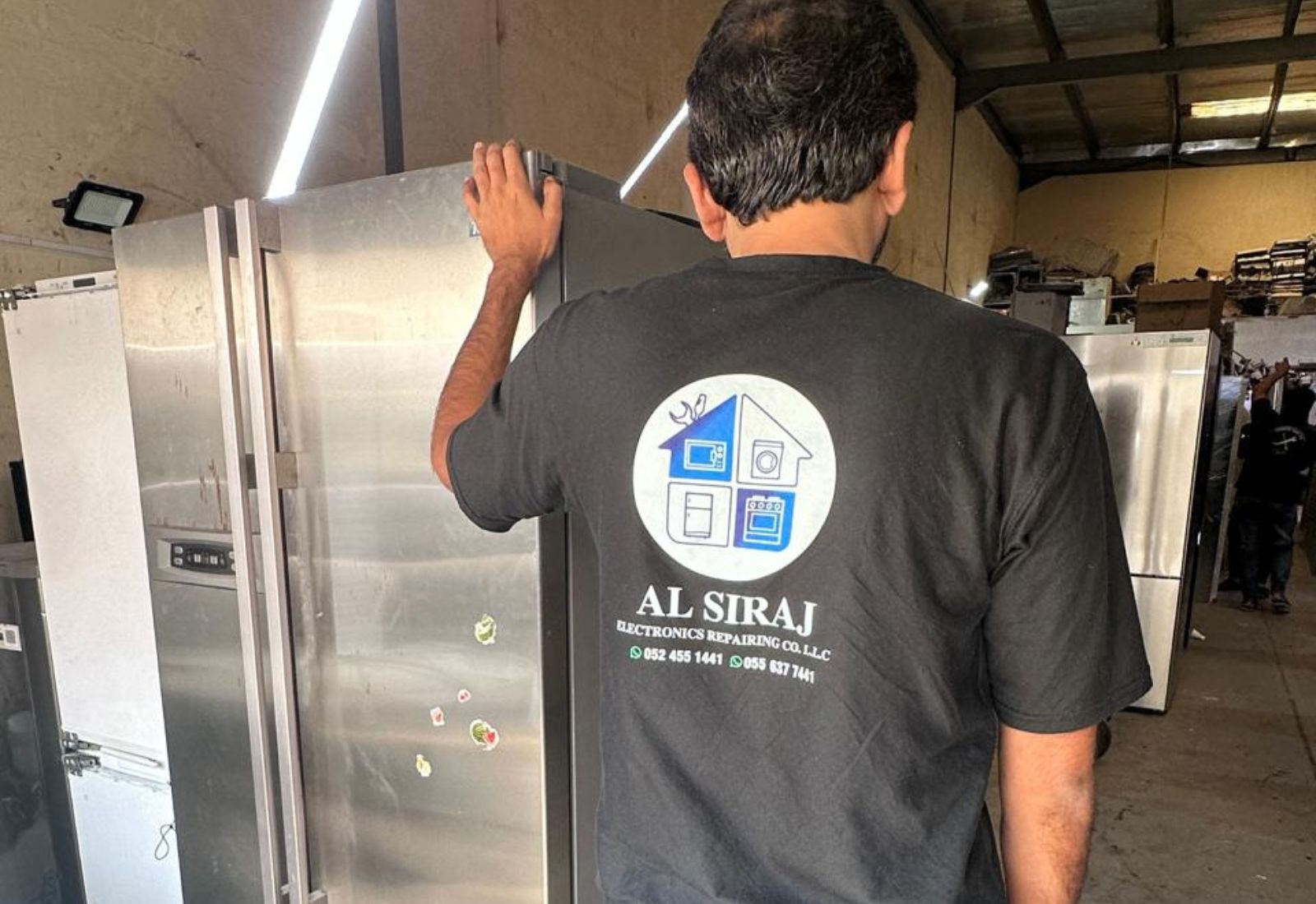 Refrigerator repair in Dubai with technician checking a large stainless steel fridge in an appliance workshop.